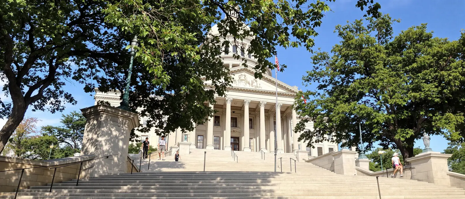 The front steps and columns of a state capitol building beside a line of mature trees