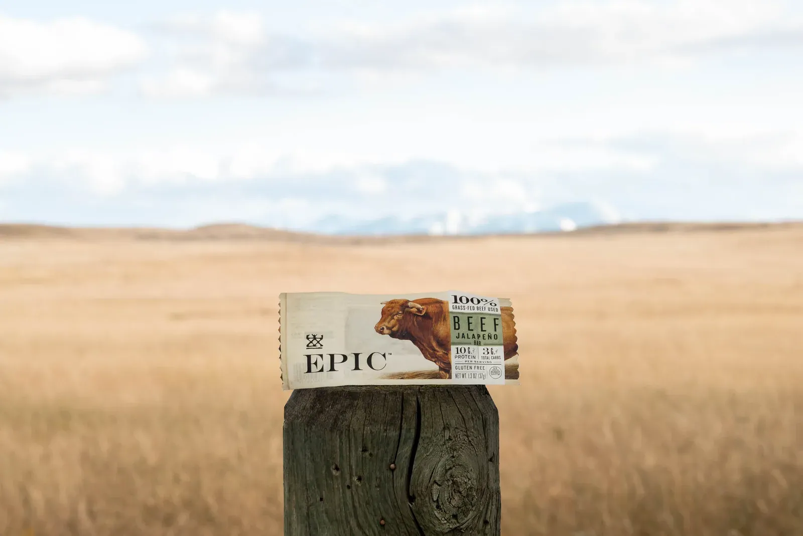 An EPIC Beef Jalapeno bar resting on a wooden fence post in front of open golden grassland and distant mountains.