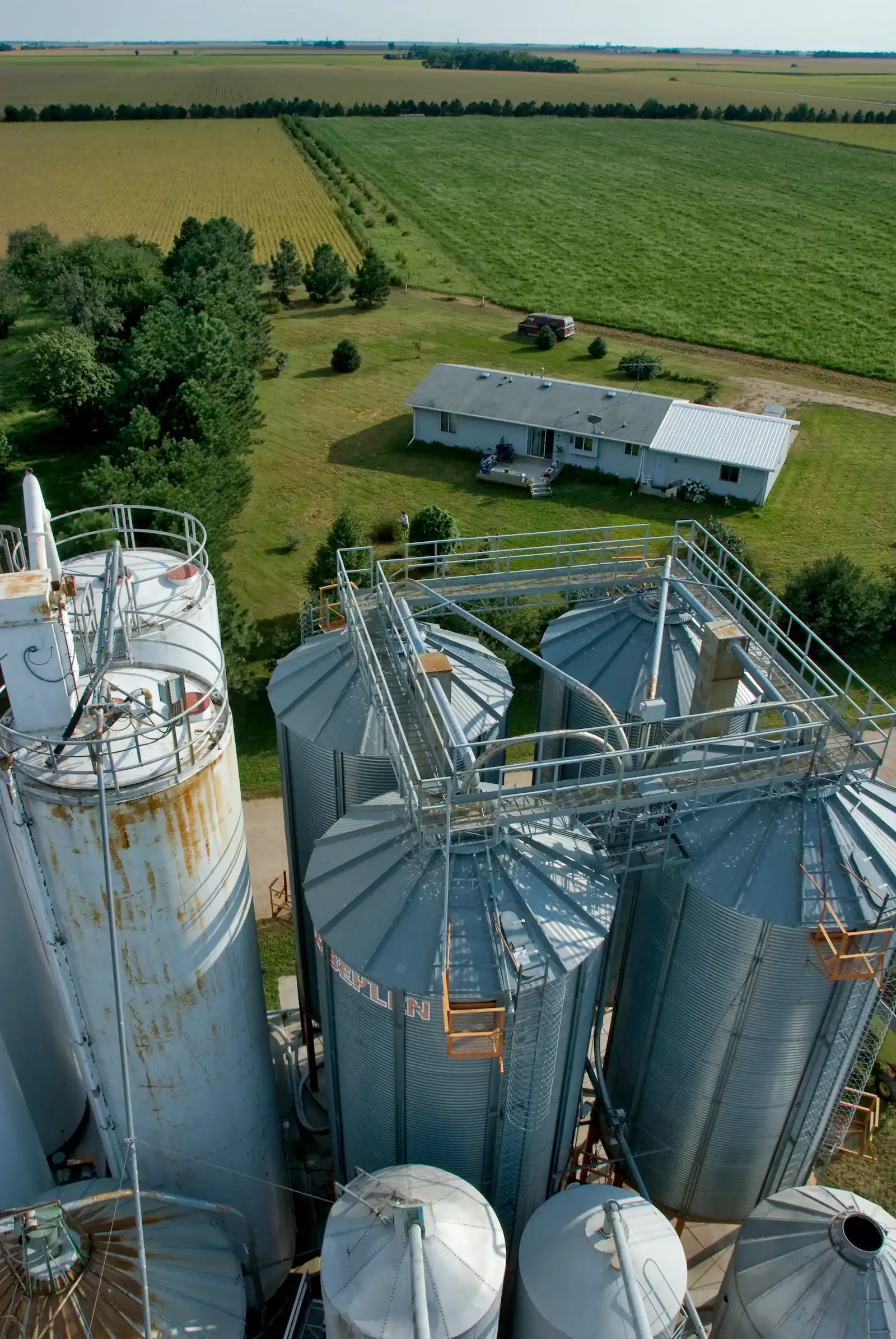 Aerial view over the grain bins of Grain Place Foods toward a farmhouse, tree rows and crop and pasture fields.
