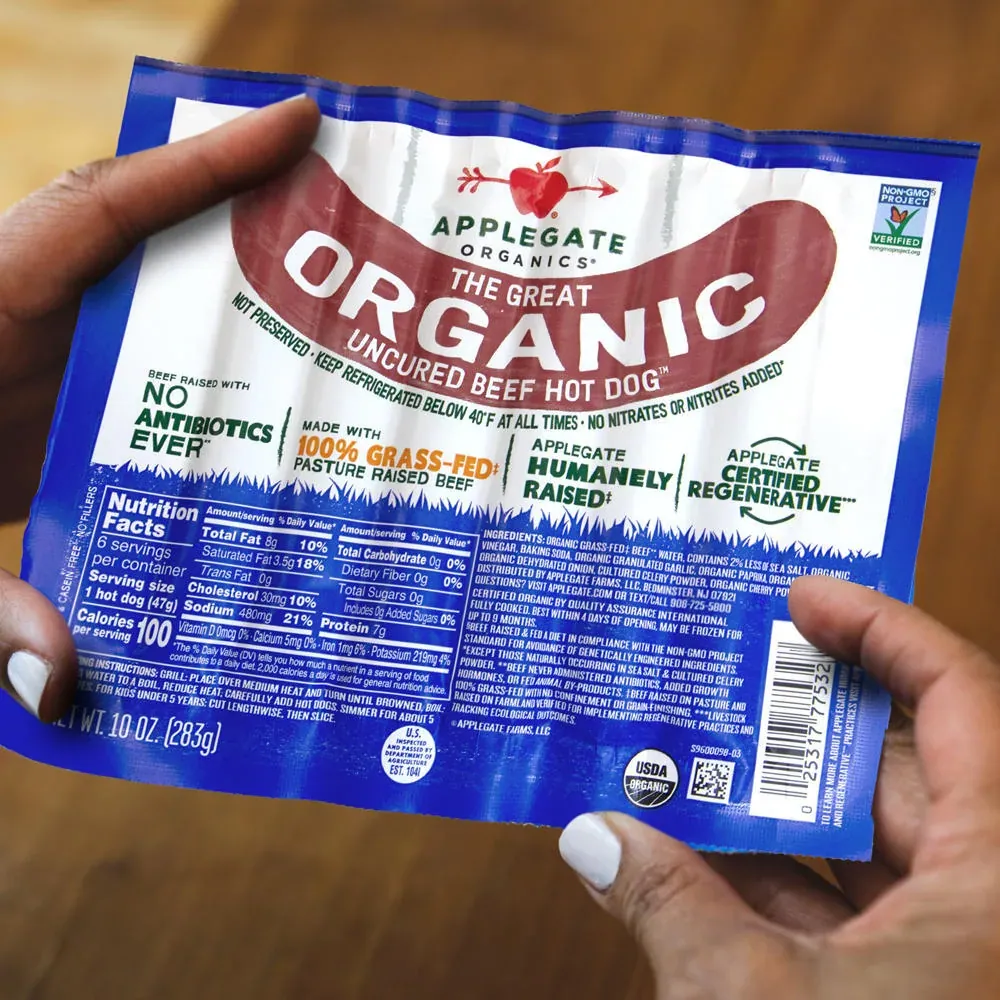 Applegate: Hands holding a pack of Applegate Organics The Great Organic Uncured Beef Hot Dog above a wooden table.