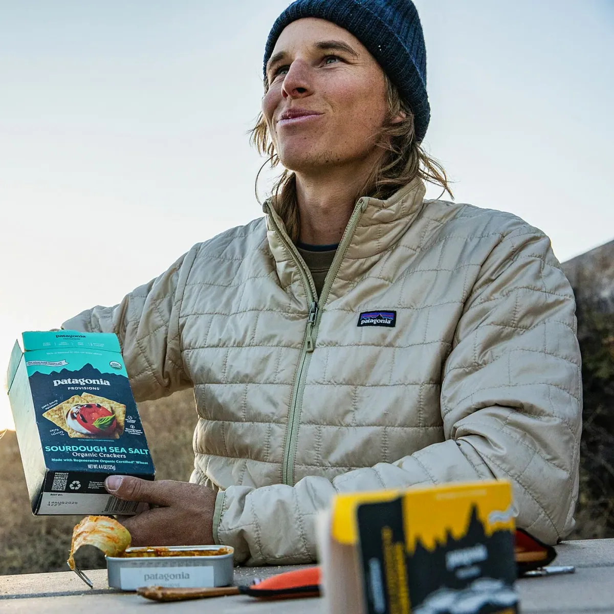 Patagonia Provisions: A person in a Patagonia jacket at an outdoor picnic table holding a box of Patagonia Provisions Sourdough Sea Salt organic crackers next to an open tin of fish.