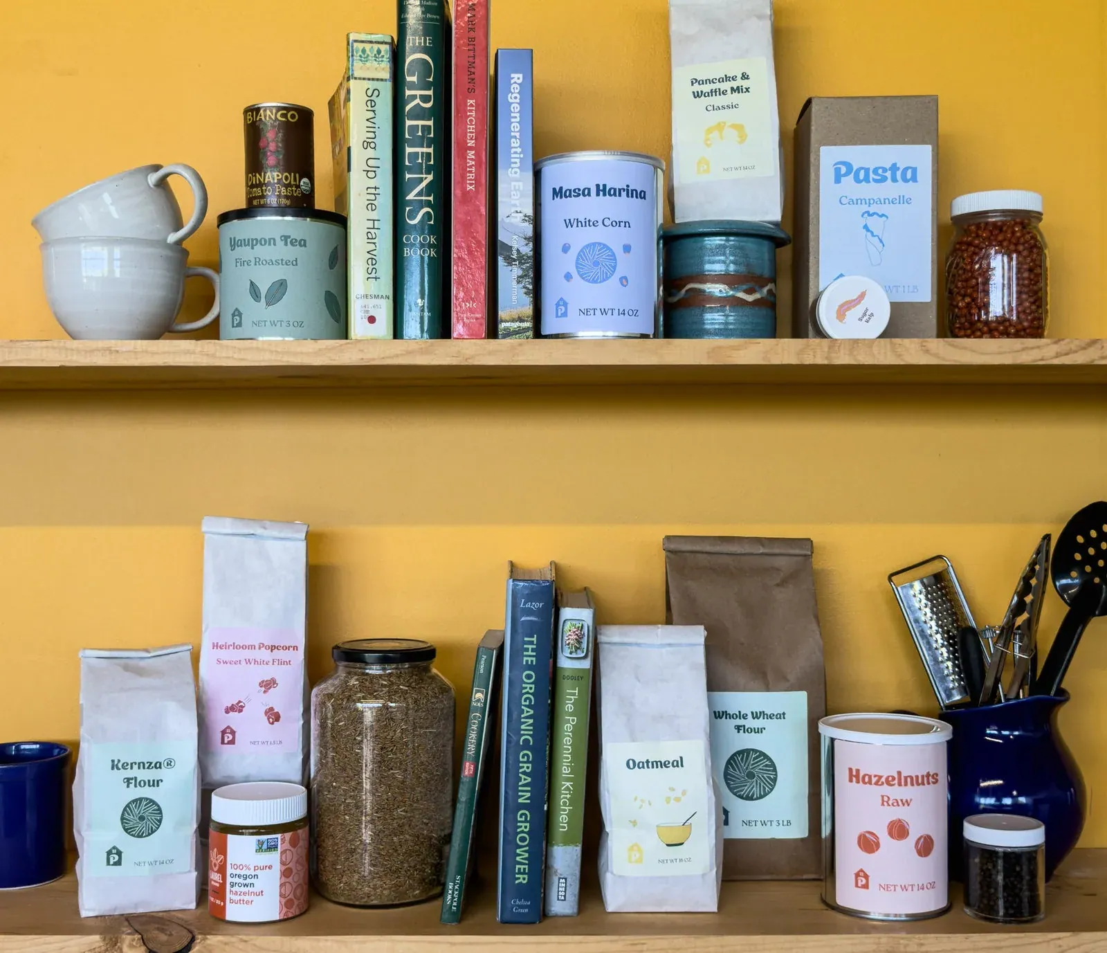 Perennial Pantry: Perennial Pantry house-label pantry goods, including Kernza flour, masa harina, oatmeal, pasta and hazelnuts, arranged on wooden shelves against a yellow wall with cookbooks.