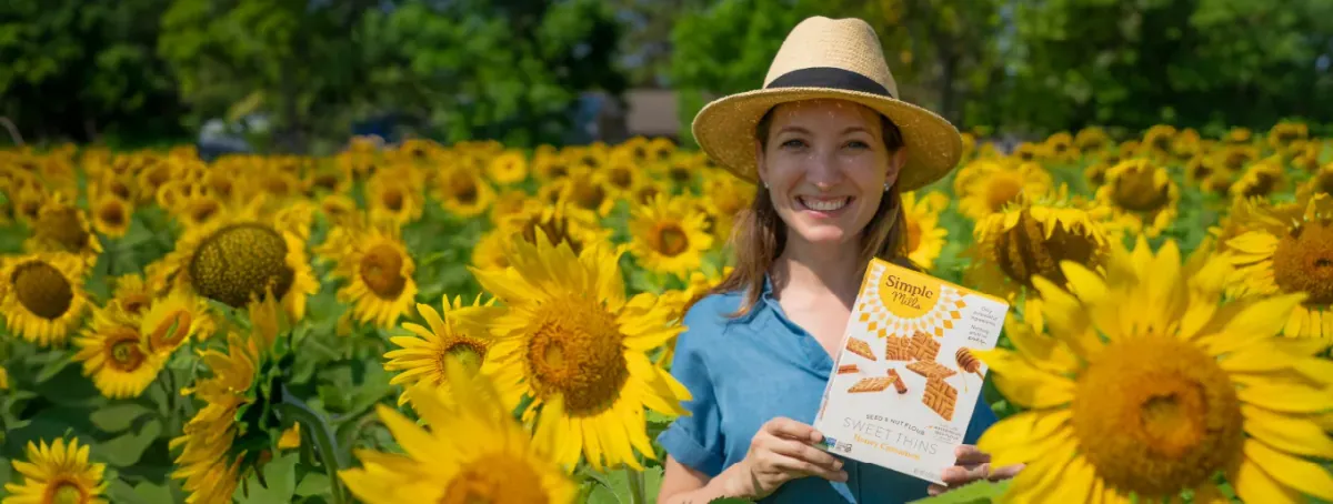 Simple Mills: Simple Mills founder Katlin Smith in a straw hat holding a box of Simple Mills crackers in a blooming sunflower field.
