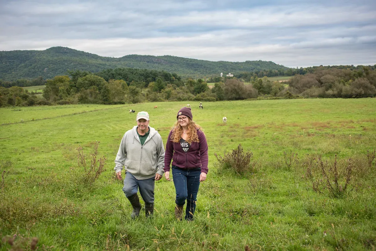 American Farmland Trust: Two farmers walk up a hillside pasture at Fuller Dairy Farm with dairy cows grazing behind them, the picture American Farmland Trust uses for its Land Transfer work on its home page.