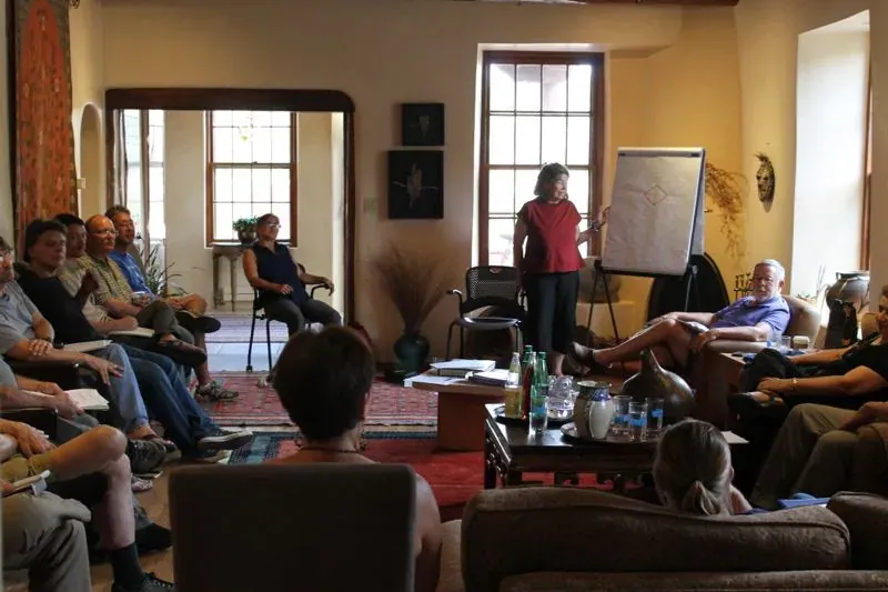 Regenesis Group: Participants seated around a living room in an adobe building listen to a facilitator at a flip chart during a Regenesis education session.