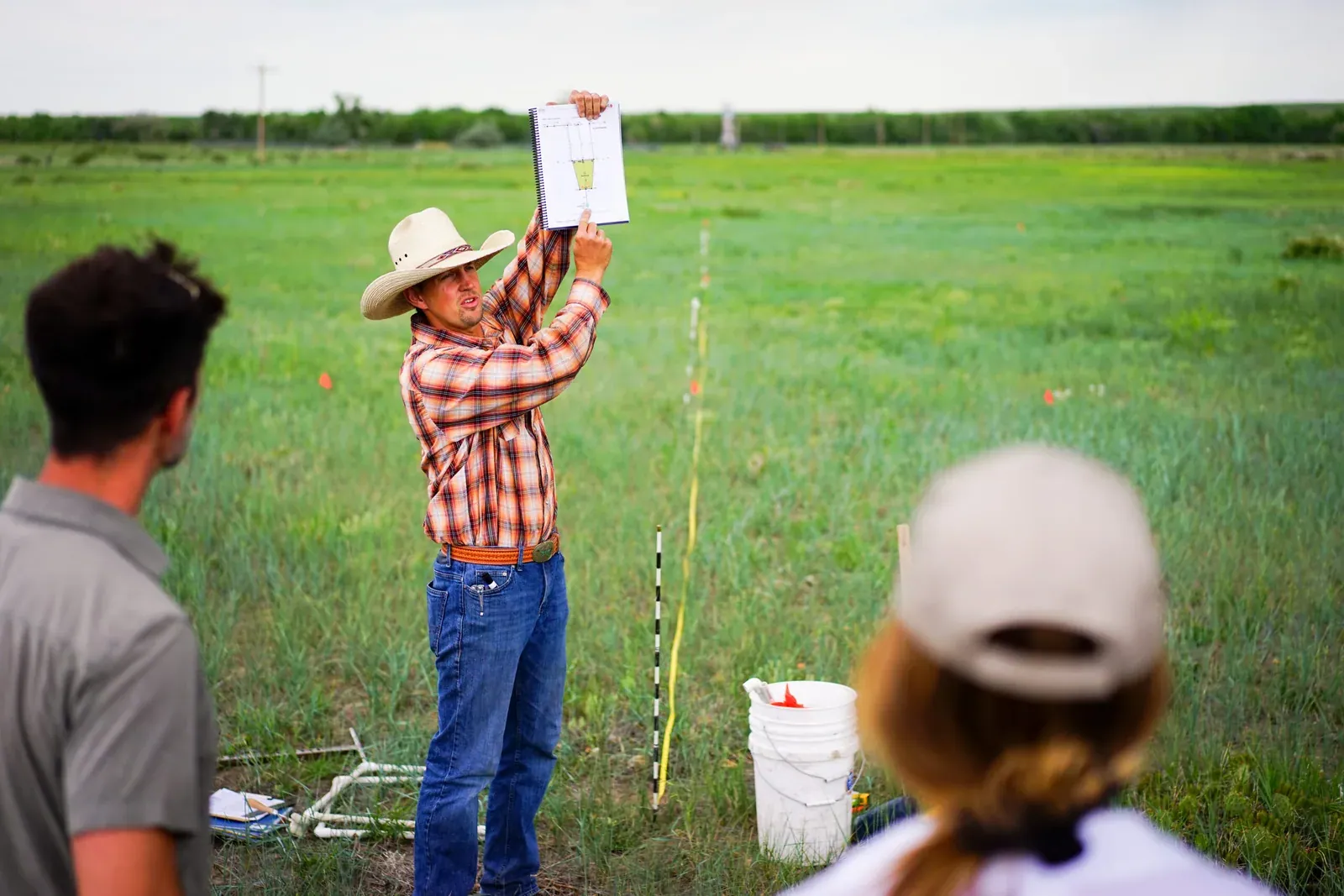 Savory Institute: An Ecological Outcome Verification (EOV) verifier holds up a clipboard in a pasture while trainees watch, with monitoring transect markers and equipment on the ground.