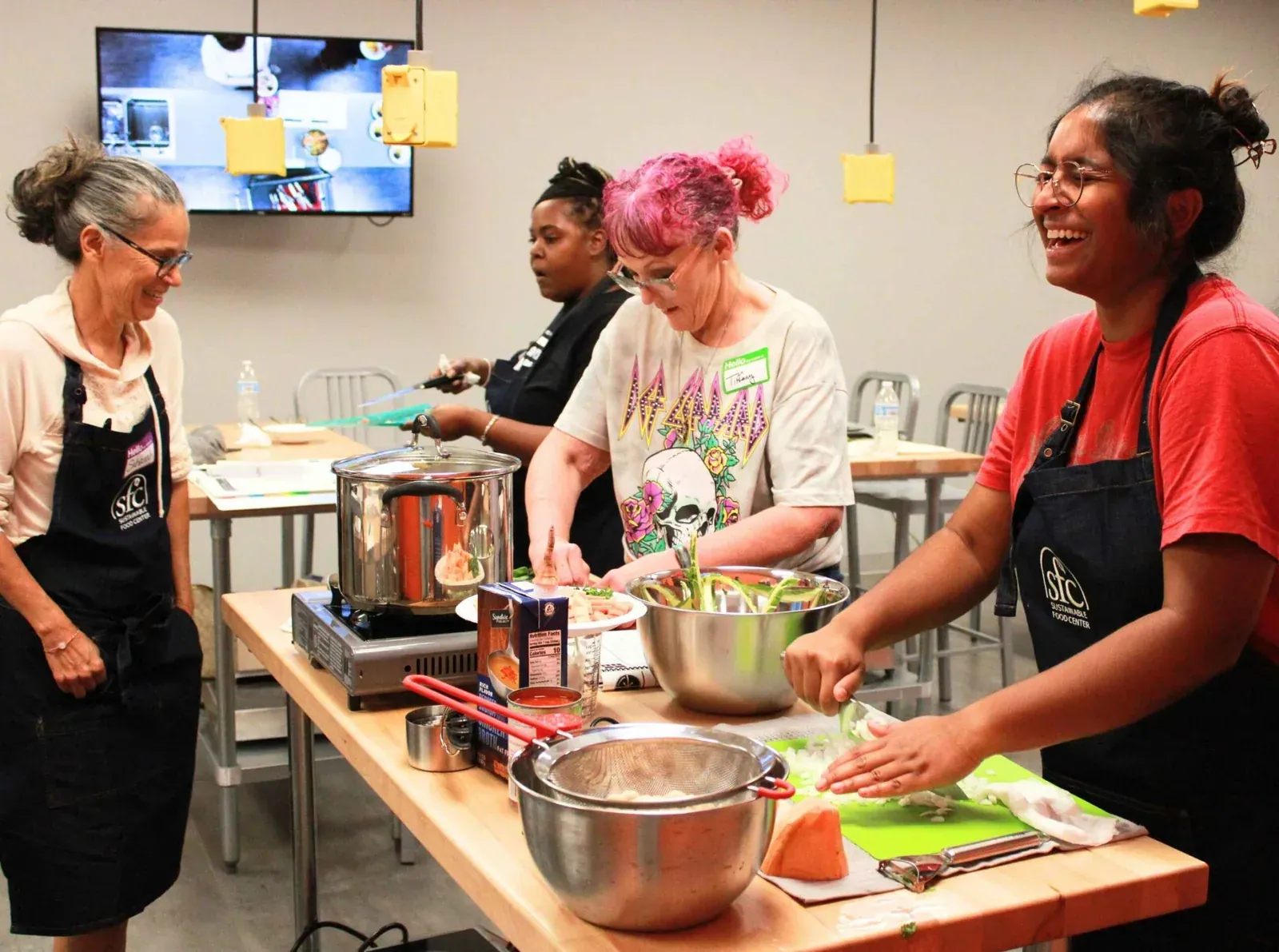 Sustainable Food Center: Participants in SFC aprons laugh while chopping vegetables at a cooking station during a Happy Kitchen / La Cocina Alegre cooking class.