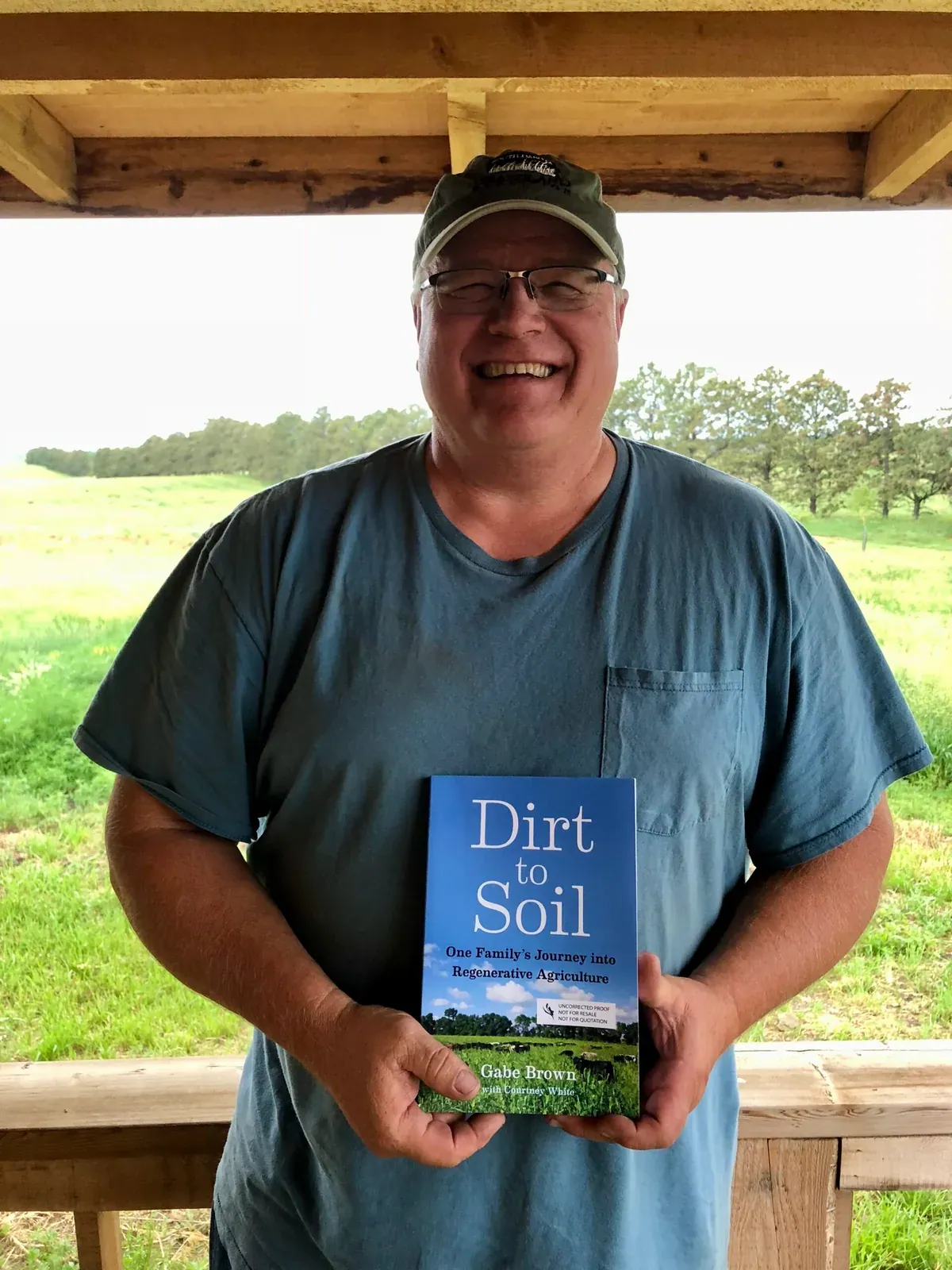 Gabe Brown: Gabe Brown in a cap and blue T-shirt holding a proof copy of his book Dirt to Soil under a shelter on the ranch, pasture and shelterbelt behind him.