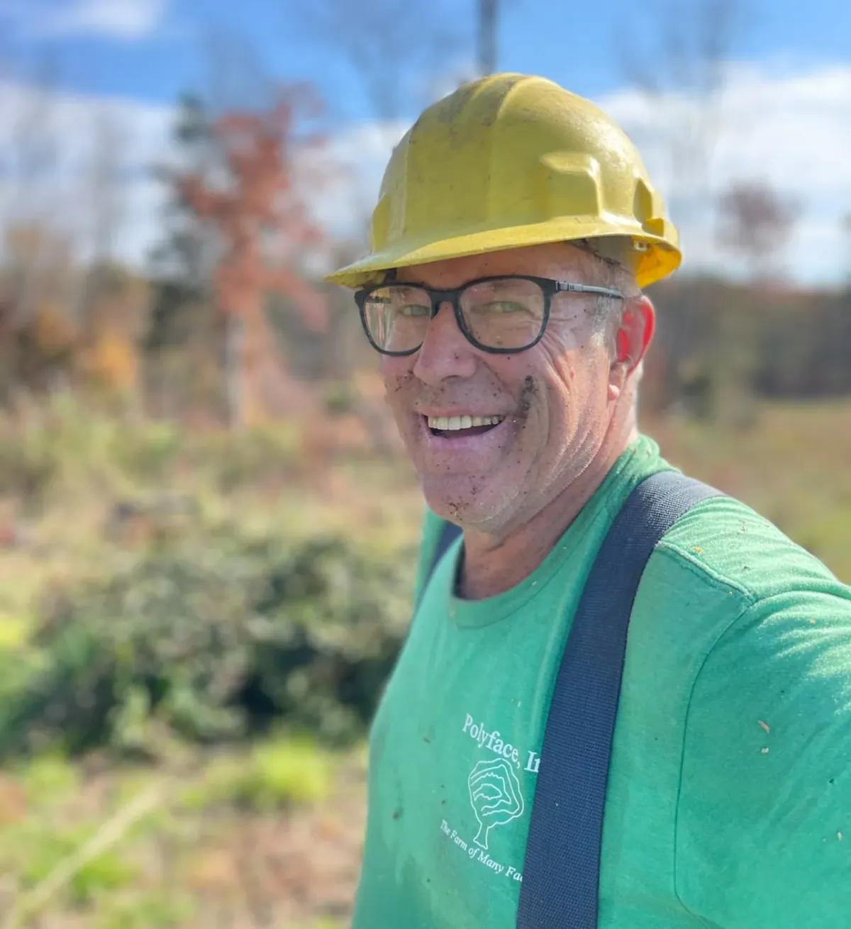 Joel Salatin: Joel Salatin smiling in a yellow hard hat, glasses and a green Polyface T-shirt, his face smudged with dirt, autumn woodland behind him.