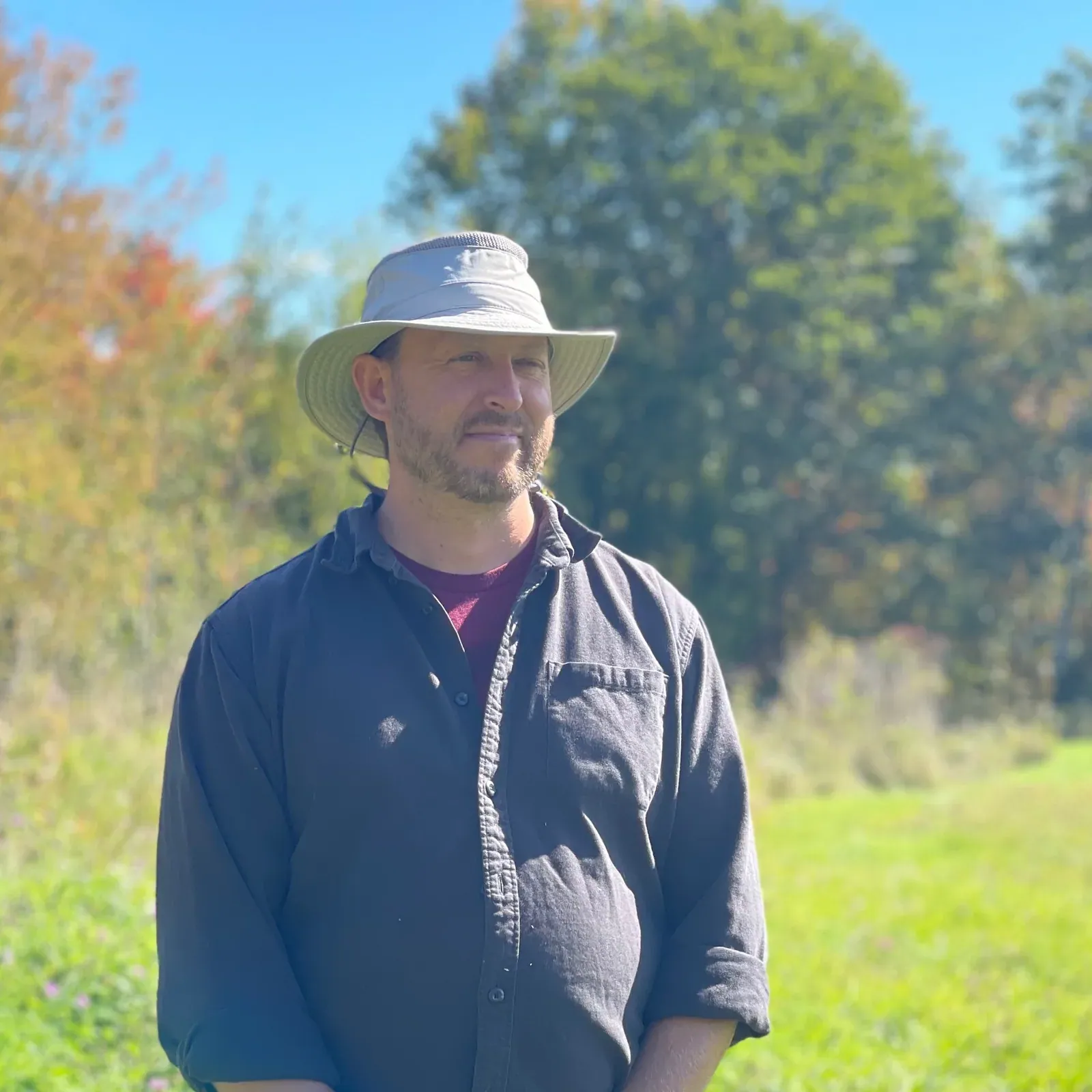 Jonathan Lundgren: Jonathan Lundgren in a bucket hat and dark shirt standing in a sunny prairie meadow with autumn trees behind.