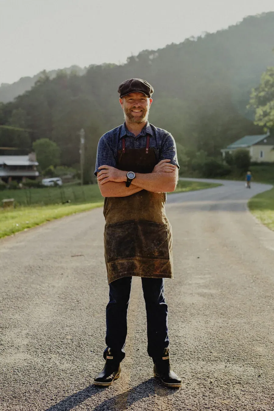 Justin Rhodes: Justin Rhodes in a flat cap and waxed work apron standing arms crossed on the gravel lane of the homestead, wooded hills behind.
