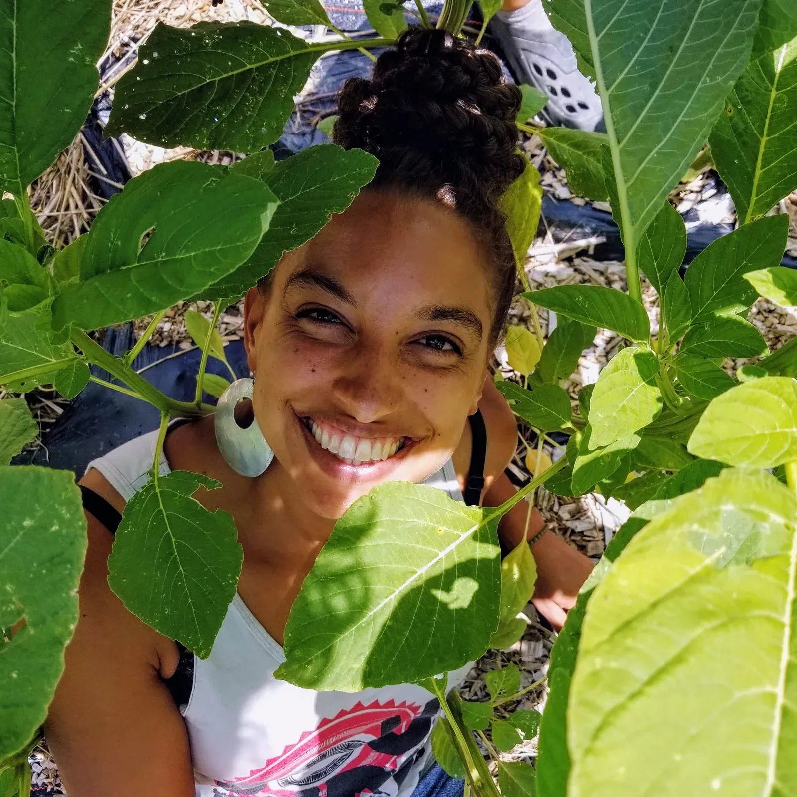 Leah Penniman: Leah Penniman smiling up at the camera from among tall amaranth leaves in a mulched garden bed.