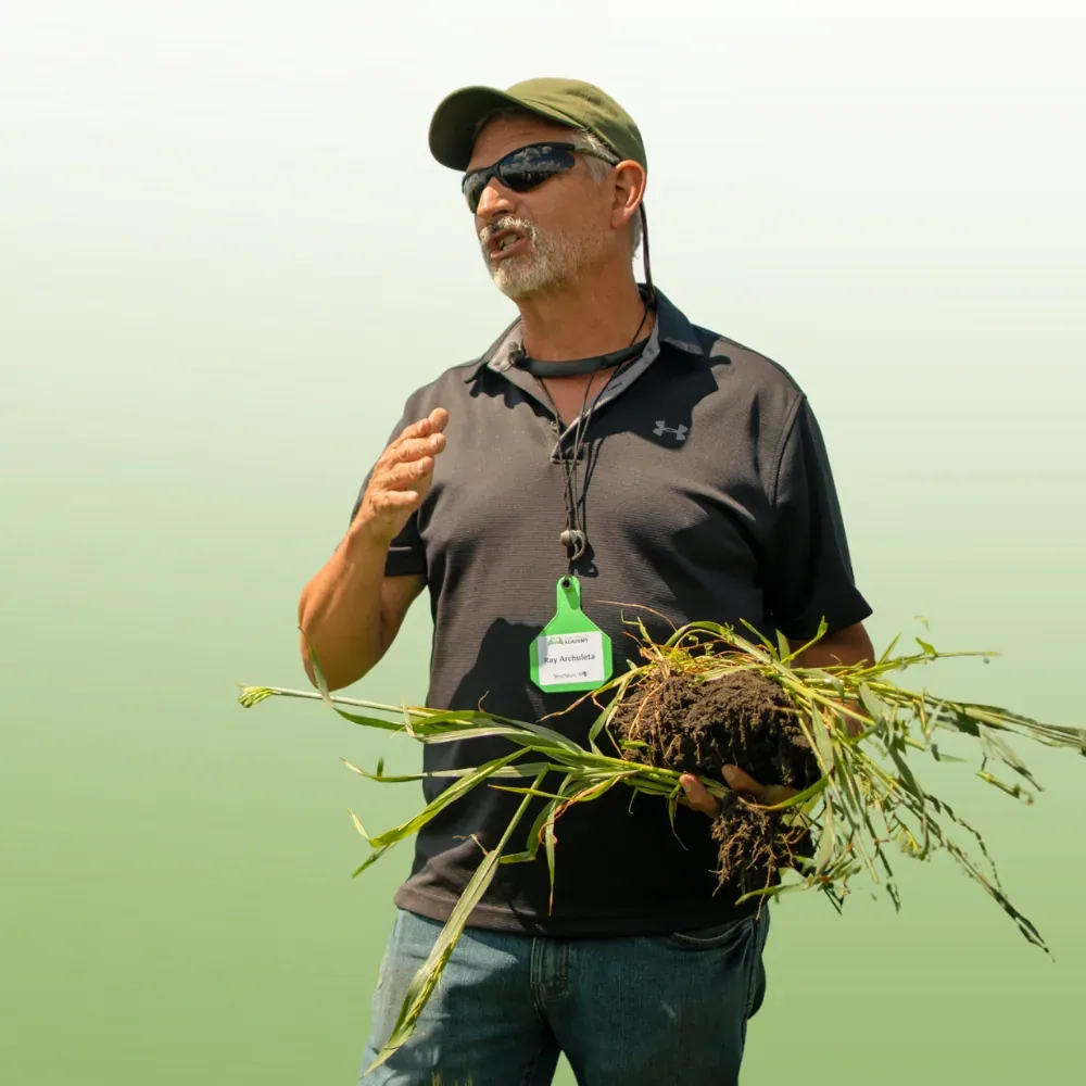 Ray Archuleta: Ray Archuleta teaching outdoors, holding a cover-crop plant with its root ball of soil, wearing a cap, sunglasses and an event name badge.
