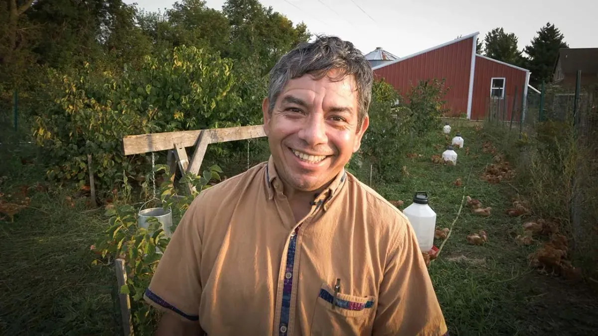 Reginaldo Haslett-Marroquin: Reginaldo Haslett-Marroquin smiling in a chicken paddock among shrubs and trees, hens and waterers around him and a red barn behind.