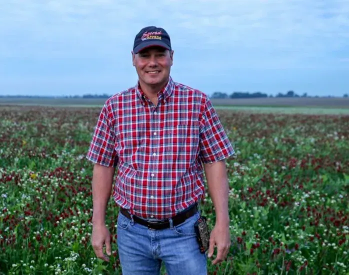 Rick Clark: Rick Clark in a plaid shirt and cap standing in a flowering clover cover crop at dusk.