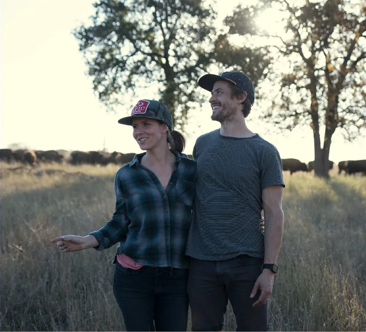 Taylor Collins and Katie Forrest: Taylor Collins and Katie Forrest standing together in tall grass with a bison herd and oak trees behind them; the right half of the file is a blank paper-texture panel that held the bio text.