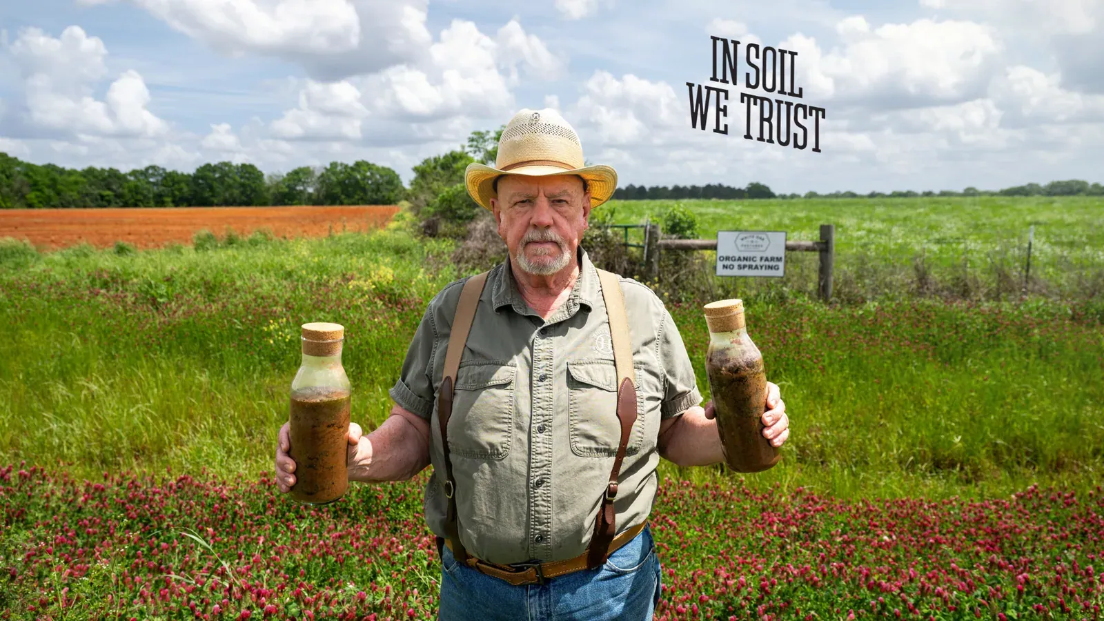 Will Harris: Will Harris in a straw hat and suspenders standing in a crimson-clover pasture holding two jars of soil, an "Organic Farm, No Spraying" sign behind him; the farm's "In Soil We Trust" headline is set into the sky at upper right.