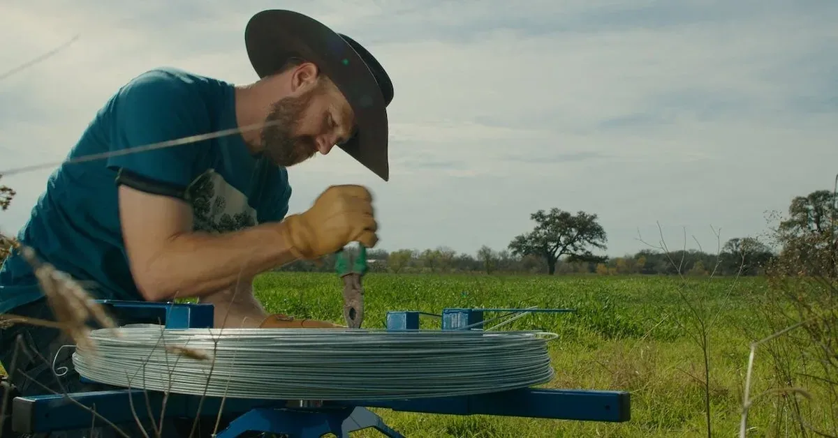 Powerflex Supply: A still from Powerflex's brand video: a man in work gloves handles a coil of high-tensile fence wire on a spinning jenny at the edge of a green pasture.
