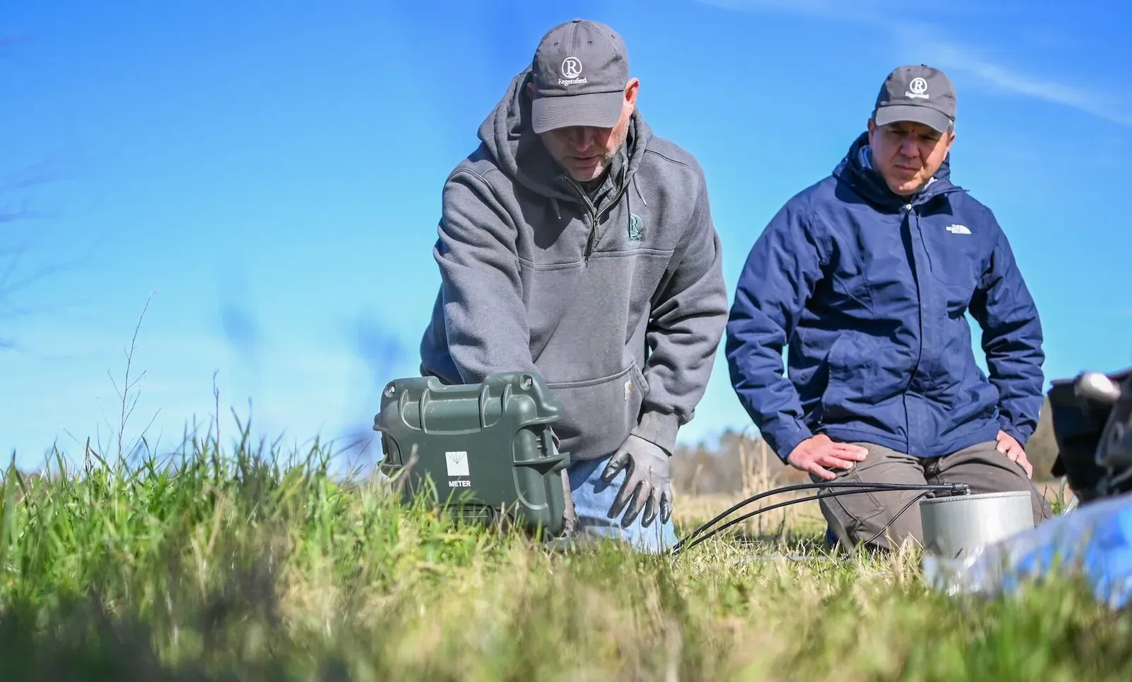 Regenified: Two Regenified verifiers in Regenified caps kneeling in a pasture beside a soil sampling case, gathering soil data in the field (Regenified's own caption).