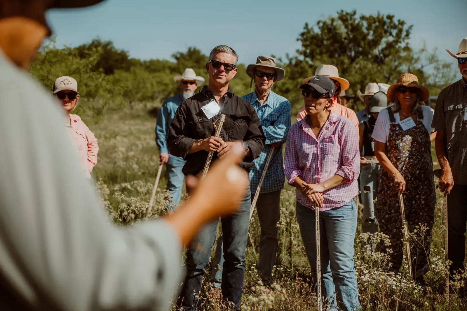 Soil Health Academy: Participants with name tags and soil probes listen to an instructor in a flowering pasture during a Soil Health Academy school in Throckmorton, Texas (May 2025).